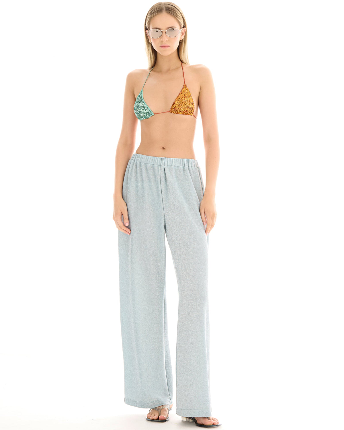 A woman wearing pale ice blue wide-leg lurex pants with a metallic shimmer, styled with a colourful triangle bikini top and flat sandals.
