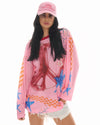 Front lifestyle view of the pink slogan trucker cap styled with sunglasses, a colourful oversized mesh top and white shorts.