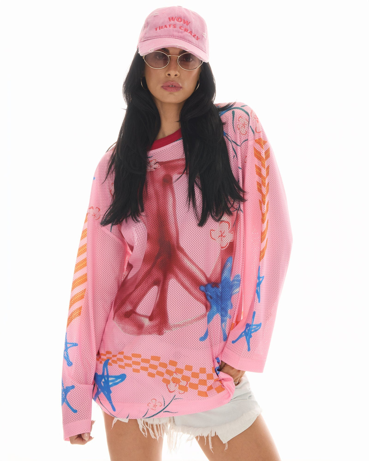 Front lifestyle view of the pink slogan trucker cap styled with sunglasses, a colourful oversized mesh top and white shorts.
